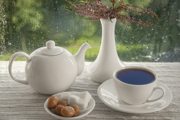 English 

black tea in a white Cup. Tea poured from a kettle. On the table, sugar, caramel and a bouquet of 

Heather in a vase. On the street it is raining. Rain drops on glass.