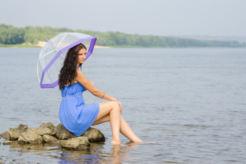 Naklejka premium Lonely sad young woman with an umbrella sits on a rock by the river