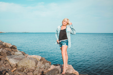 Tanned woman posing at the beach on summer day, looking up. Dreamy female in jeans clothes, standing rocky coast, blue sky background with sea. Concept holiday, outdoors. Travel active lifestyle.