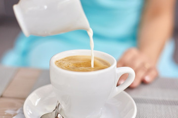 Lady in blue dress pours milk in white cup with coffee