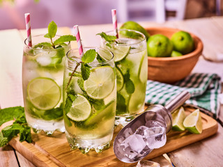Alcohol cocktail. On wooden boards three glasses with alcohol cocktail and ice cubes. Drink number hundred and ninety two cocktail mohito with straw and lime and mint leaf. Country life. Indoor.