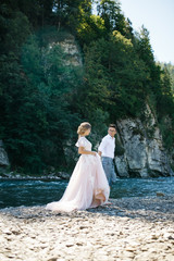 Groom holds bride's hand while walking on the river's shore