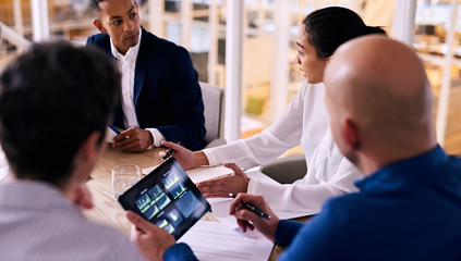 Group of four business people talking to each other during a business meeting with a caucasian man holding an electronic tablet in his hands with graphs and charts on the display.