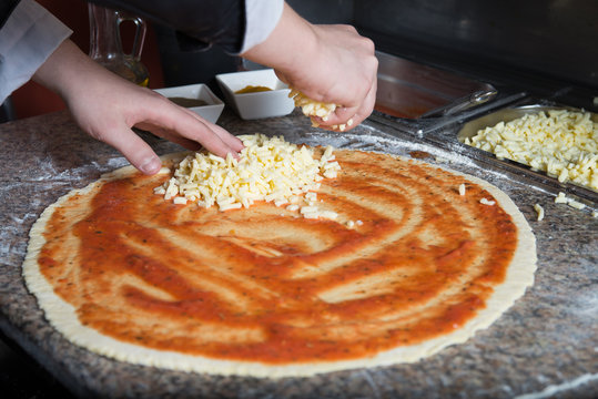 Chef sprinkling mozarella cheese over the top of pizza dough. He makes it by own hands. At worktop lie aromatic spices, tomato sauce and mozarella cheese.