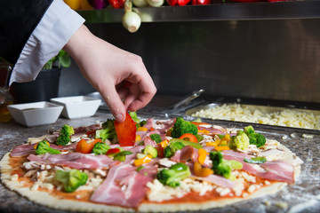 Preparating traditional pizza in profesional pizzeria. Chef is covered pastry by tomato sauce, mozarella cheese, mushrooms, bacon and fresh vegetables like pepper and broccoli. 