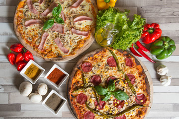 Two pizzas at wooden background in the company of fresh ingredients like spices, vegetables and herbs.