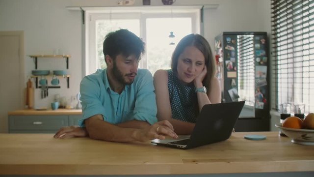 Young good-looking couple is sitting at the kitchen table looking at the computer and talking. Slow motion