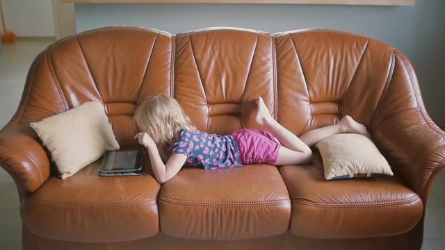 A side view of a cute blond little girl wearing all pink is lying on the sofa and playing a game on tablet. Slow motion