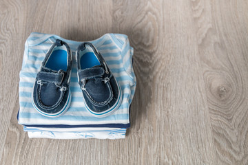 Folded blue and white bodysuit with shoes on it on grey wooden background. diaper for newborn boy. Stack of infant clothing. Child outfit. Copy space.