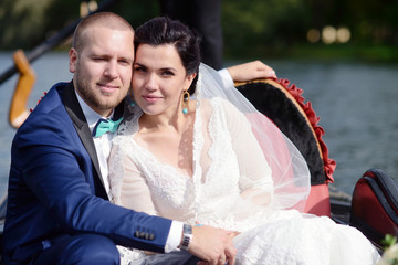 Wedding couple is hugging on a floating gondola. Beauty bride with groom. Beautiful model girl in white dress. Man in suit. Female and male portrait. Woman with lace veil. Cute lady and guy outdoors