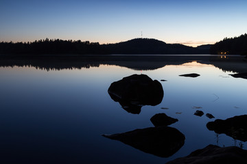 Silhouettes and reflections of a forest and rocks at a calm lake in Finland in summertime at dusk.