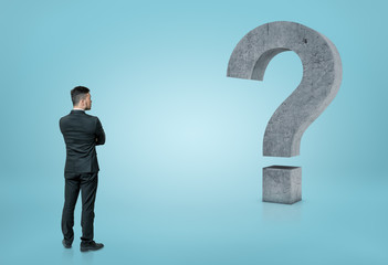 Back view of a businessman looking at big 3D concrete question mark isolated on blue background
