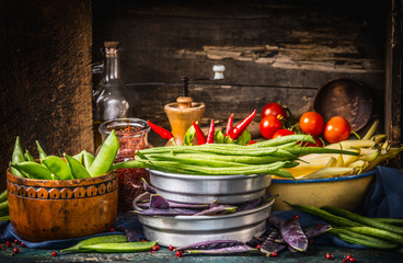 Various colorful pods peas and beans in bowls with cooking ingredients on dark rustic kitchen table with wooden background