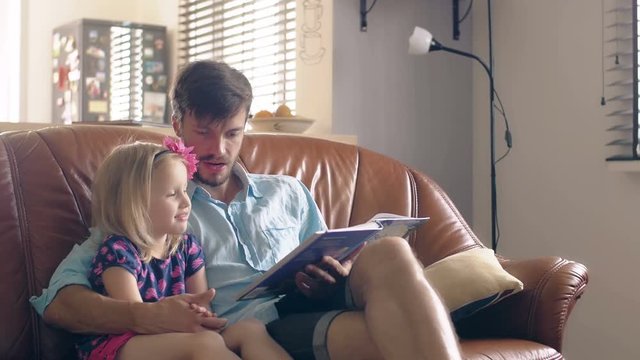 A young father and his happy little blond daughter are reading a story on leather sofa in the dining room. 4K