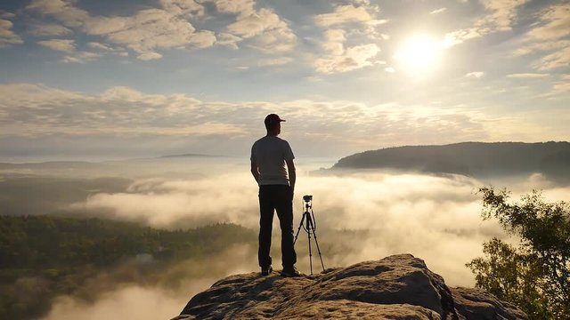 Photographer in grey t-shirt and red cap preparing tripod on cliff. Dreamy fogy landscape, blue misty sunrise in a beautiful valley below