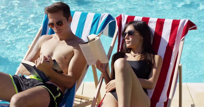Young Caucasian and Asian male and female couple in sunglasses reading books on deck chairs at the swimming pool