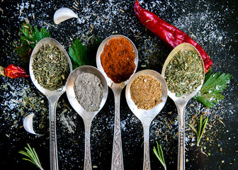 Spices in spoons on a black background, with vegetables and herbs