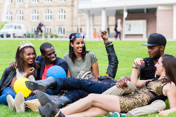 Multikulturelle Gruppe Männer und Frauen sitzt zusammen auf Rasen und bläst Seifenblasen