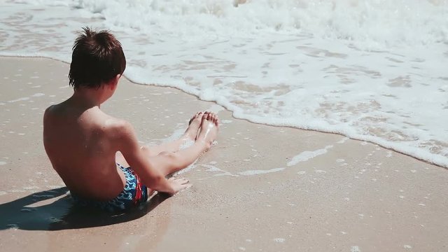 Boy 9 years old sitting on the sand of the sea by the sea. Playing sea sand, waves wash the feet of the child.