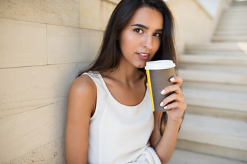 Beautiful close-up portrait of young attractive brunette woman with a cup of coffee or tea, sitting on the steps in the park. Looking at the camera.