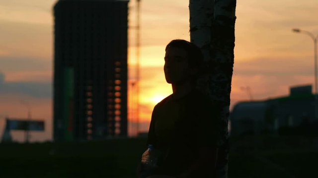Athlete man resting at sunset and drinks water from a bottle. Sun beautifully reflected in the water. City landscape with cars and construction site