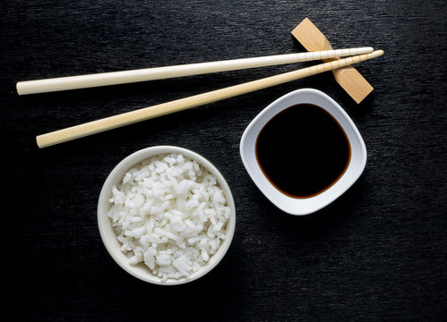 Japanese sushi chopsticks over soy sauce bowl, rice on black bac