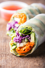 fresh and vegan rice paper spring rolls with raw vegetables (cucumber, avocado, lettuce, carrots, red cabbage) inside, sweet chili dipping sauce behind, isolated over dark, wooden background