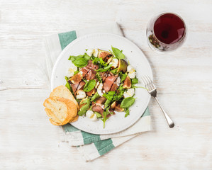 Prosciutto, arugula, basil, figs and mozzarella salad with glass of red wine over white wooden background, top view