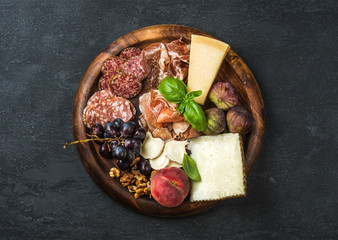 Italian antipasti snack for wine. Prosciutto di Parma, salami, cheese variety, figs, grapes, peach, walnuts and fresh basil on wooden serving tray over dark grunge background. Top view