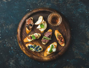 Italian crostini with various toppings and glass of wine on round wooden serving tray over black plywood background, top view