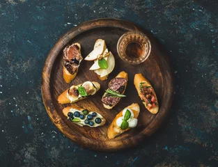 Zelfklevend Fotobehang Italian crostini with various toppings and glass of wine on round wooden serving tray over black plywood background, top view © sonyakamoz