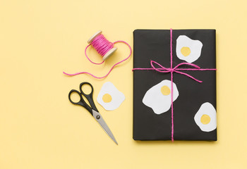 Top view on cute present wrapped in black paper and decorated with eggs shapes on balck background with scissors and pink ribbon. Funny gift for birthday party or any celebration. Holidays concept.