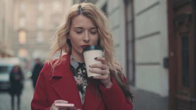 Business woman in red coat with coffee walking in the city and communicate via smart phone (phablet). Steady cam shot.