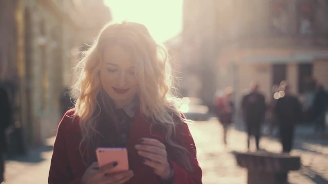 Business woman sms texting using app on smart phone. Attractive girl in red coat walking in the sunshine old city and communicate via smart phone (phablet). Steady cam shot.