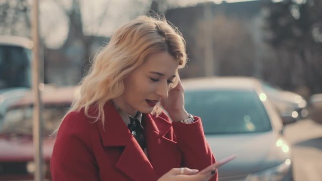 Attractive girl in red coat walking in the sunshine city and communicate via smart phone (phablet). Steady cam shot.
