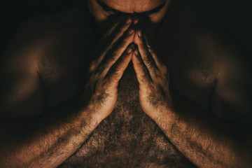 Man in despair, holding hands in prayer in front of his face, alone in a dark room