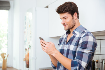 Naklejka na ściany i meble Handsome young man standing and using smartphone on the kitchen