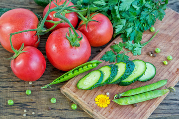Cluster of ripe red tomatoes with water drops, sliced cucumber on wooden cutting board, bunch of parsley, green peas and peppercorn on the table, fresh vegetables, salad ingredients
