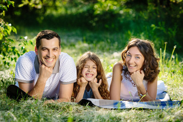Fototapeta premium Young beautiful family of three having fun together outdoor. They are lying on a lawn look happy and smile. Fun outside sunny day.
