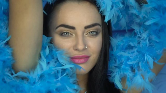 Closeup portrait of beautiful seductive brunette woman smiling and looking into the camera while playing with blue feather boa over blue wall background
