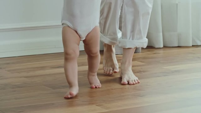 Adorable happy baby smiling while making first steps with assistance of mother 