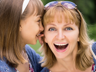 Smiling mother and daughter whispering gossip - relationships and happiness concept. A little girl telling her mother a secret. Portrait of happy family - sharing secrets, outdoors at park.