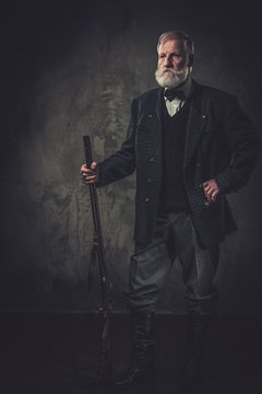Senior hunter with a shotgun in a traditional shooting clothing, posing on a dark background.
