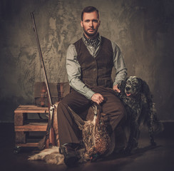 Handsome hunter with a english setter and shotgun in a traditional shooting clothing, sitting on a...