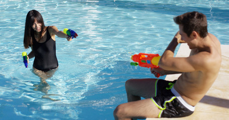 Young adults shooting shooting plastic water guns at each other in swimming pool outdoors