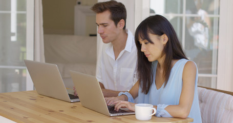 Calm young couple in loose summer clothing sitting at wooden table using similar laptop computers. Includes copy space.