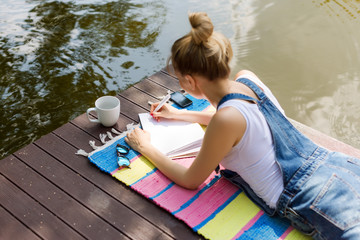 Woman making plans, taking notes in calendar or writing in her diary lying on the pier and drinking...