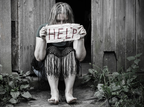 The girl sits in front of the open door, and holding a piece of paper with the word "help"