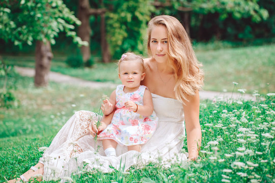 Group of two people, white Caucasian mother and baby girl child in white dress sitting playing in green summer park forest outside, looking in camera,  lifestyle happy childhood concept