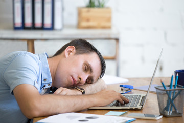 Portrait of young man lying on the table in front of laptop, sleepy, tired, overworked or lazy to work. Attractive business man napping in his home office relaxing after work on laptop computer